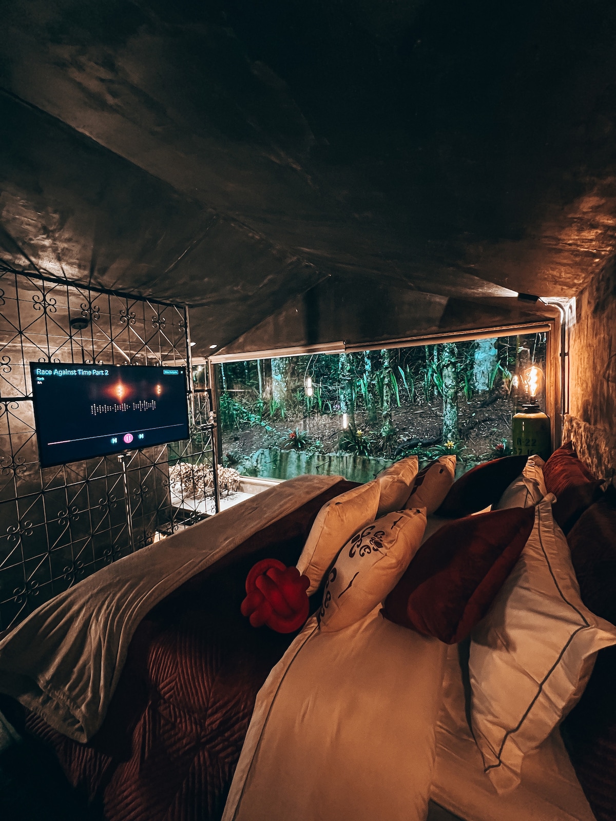 A cozy sleeping area is featured with a spacious bed surrounded by earthy tones. A large window offers views of the lush, green surroundings, while soft lighting from industrial-style fixtures enhances the atmosphere. A television screen is mounted on a wall nearby.