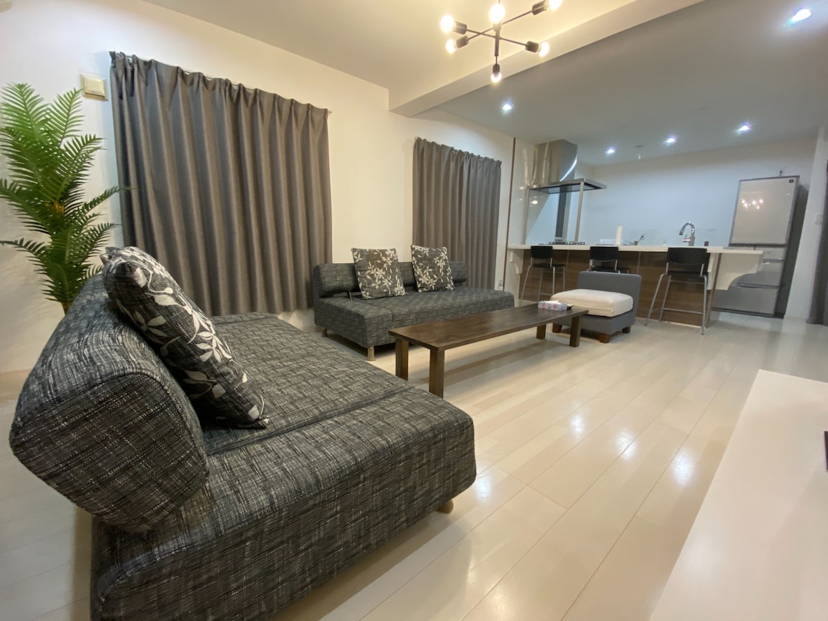 A spacious living area features a modern design with dark gray sofas adorned with patterned cushions. A wooden coffee table centers the space, while a well-equipped kitchen is visible in the background, highlighted by stainless steel appliances. Light drapes frame large windows with soft, neutral curtains.