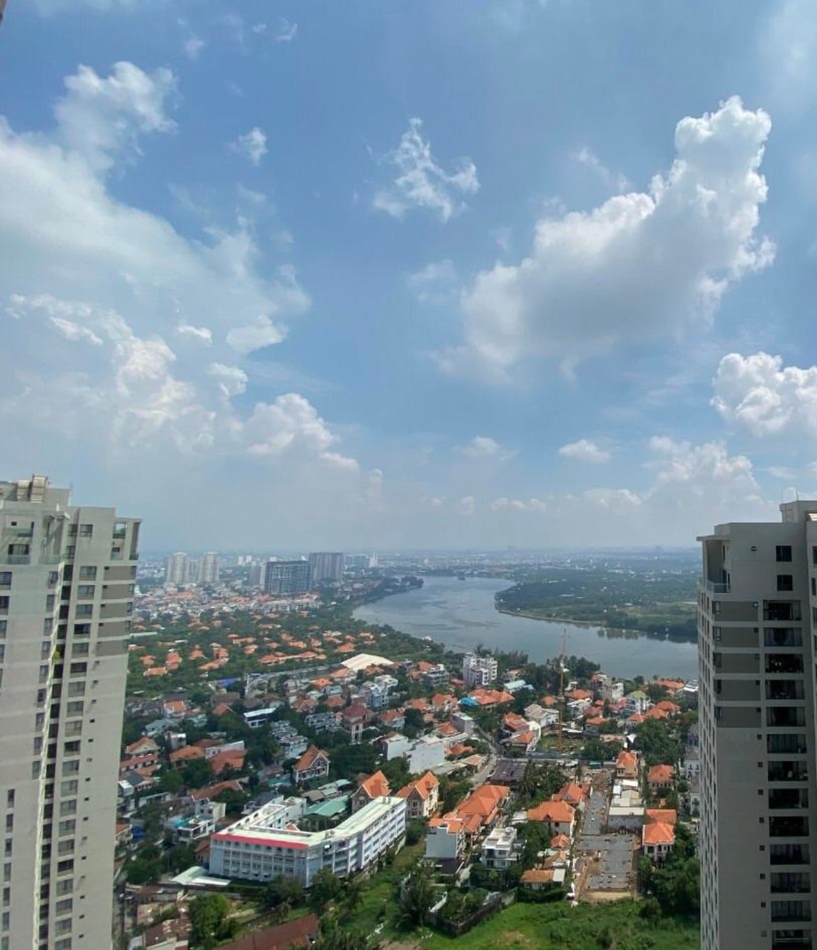 A panoramic view captures the sprawling landscape of homes with red-tiled roofs, bordered by lush greenery. A river winds through the scene, reflecting the blue sky and scattered clouds above. Tall buildings frame the horizon, showcasing the urban setting.