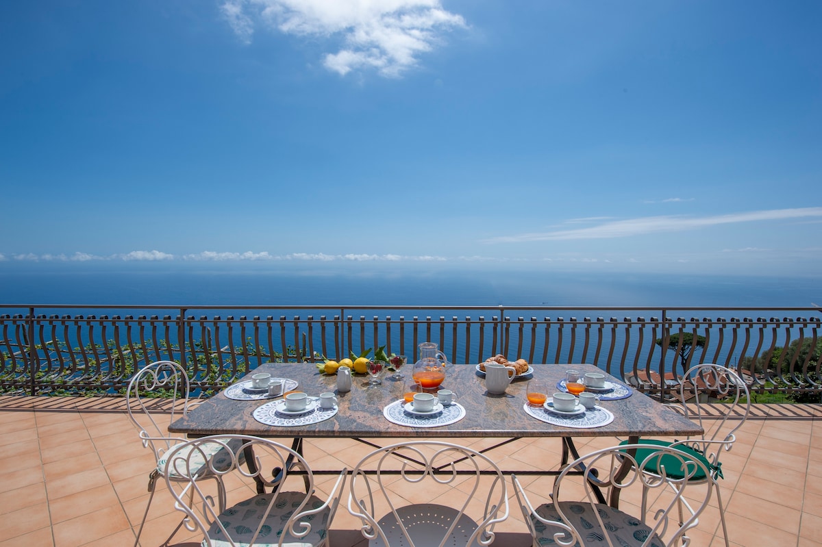 An outdoor dining area features a marble table set for six, complete with dishes and drinks. Surrounding views showcase the sea under a clear blue sky. The terrace is enclosed by wrought iron railing, providing an unobstructed perspective of the horizon.