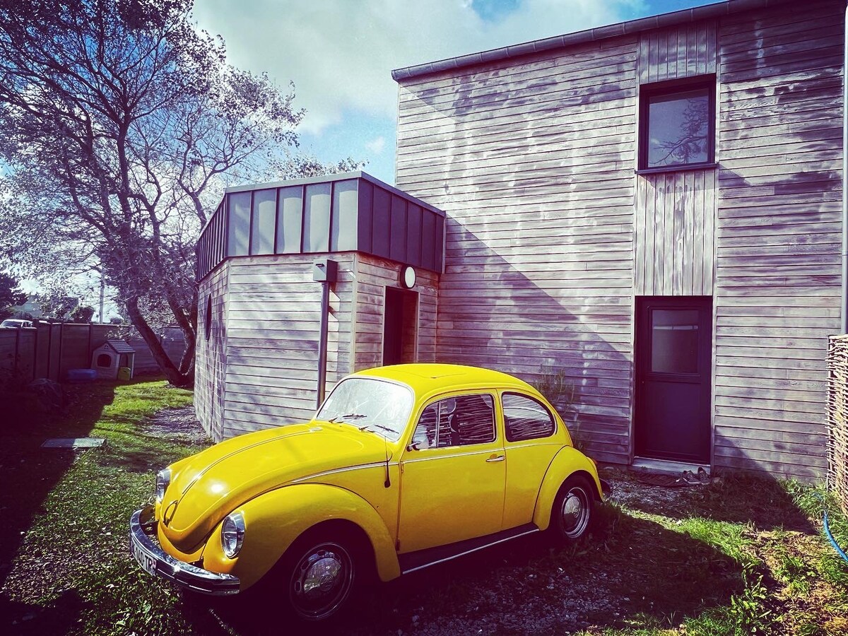 A vibrant yellow vintage car is parked near the contemporary wooden house, featuring multiple windows. The exterior is complemented by grassy areas and a tree providing shade, contributing to a relaxed outdoor environment.