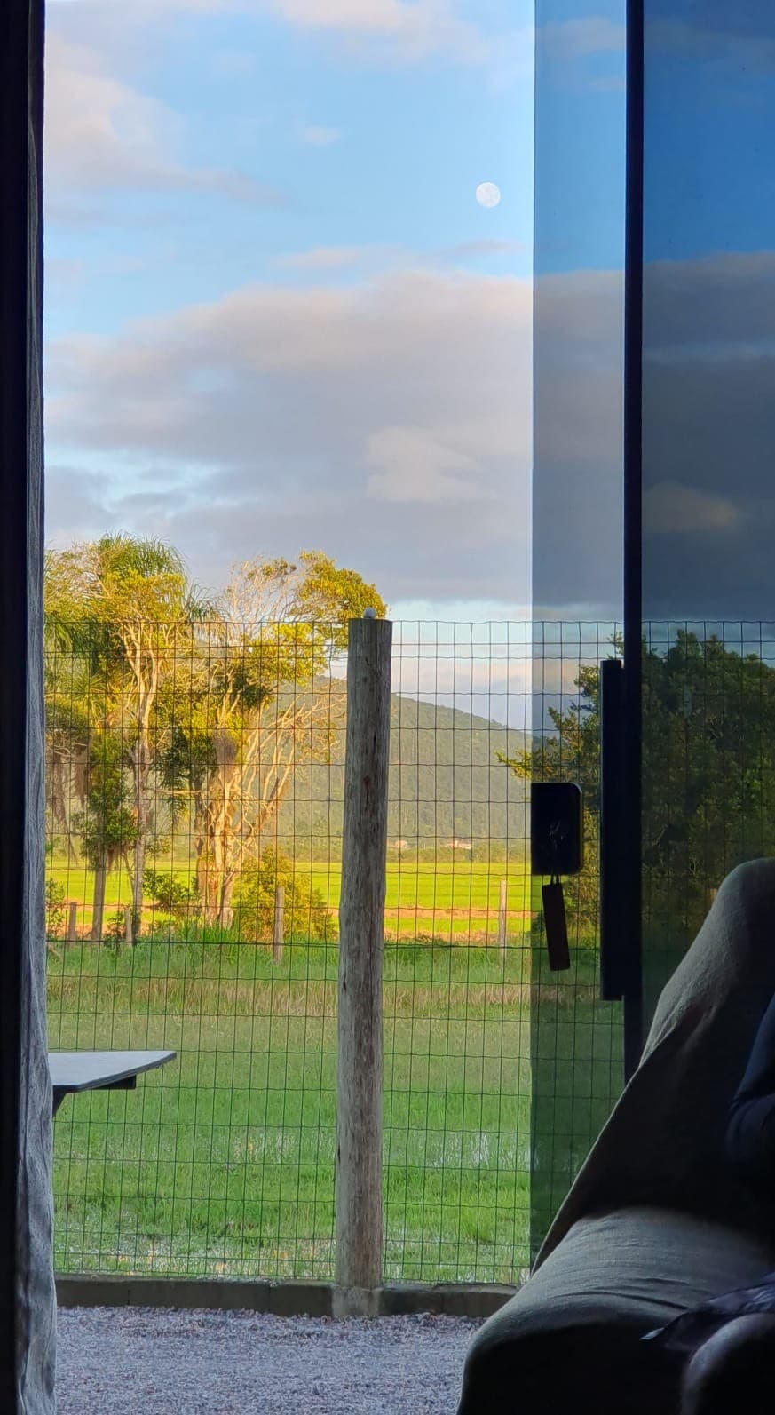 A serene view is offered from the space, featuring a fenced area with trees in the background and expansive greenery. The sky showcases a mix of clouds and a visible moon, creating a peaceful atmosphere. Natural light enters through the glass door, enhancing the open feel.