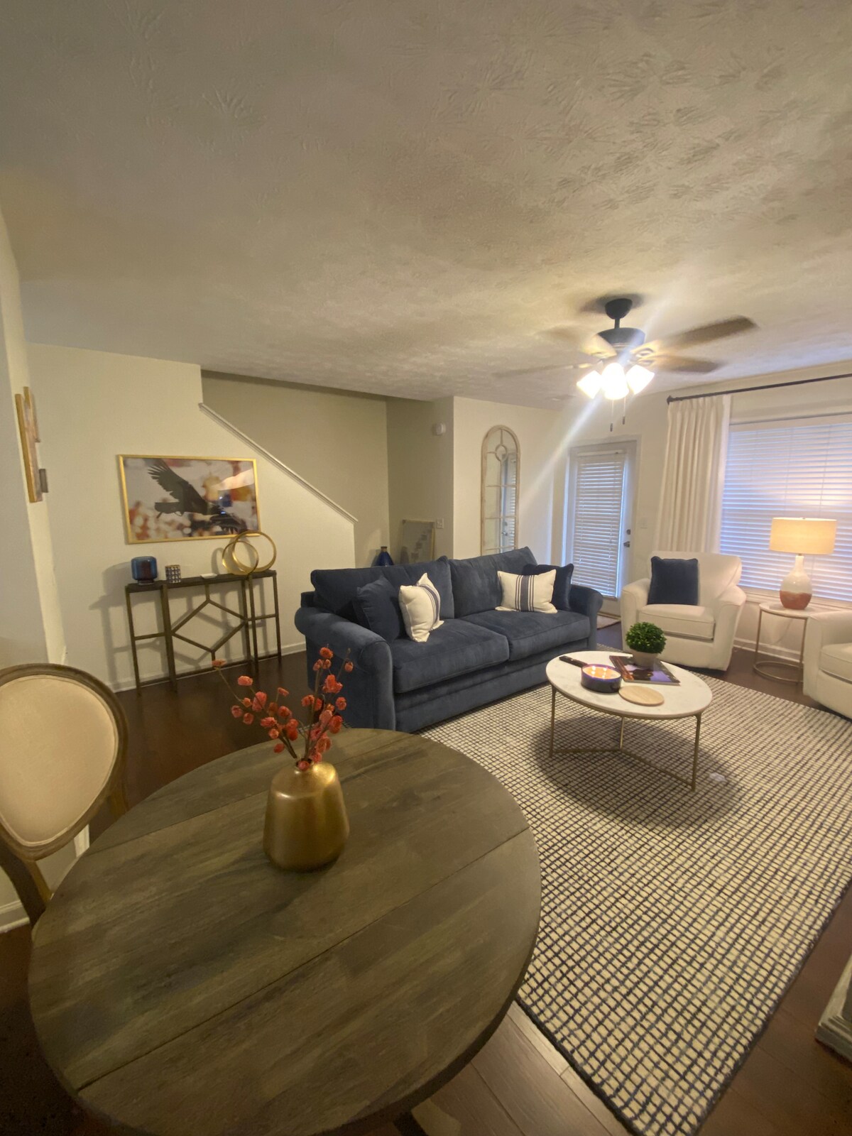 A modern living area features a blue sofa with decorative cushions, a light rug, and a circular coffee table adorned with a small potted plant. A dining table with wooden texture is visible, while natural light filters in through the windows adorned with light drapes.