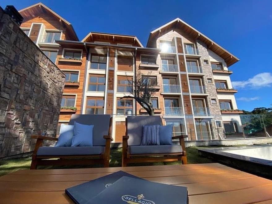 The exterior of a modern building is depicted, featuring wooden and stone accents. Comfortable seating is positioned in the foreground, with a view of expansive windows that reflect the clear blue sky. The surrounding area includes greenery and a pool, enhancing the overall serene setting.