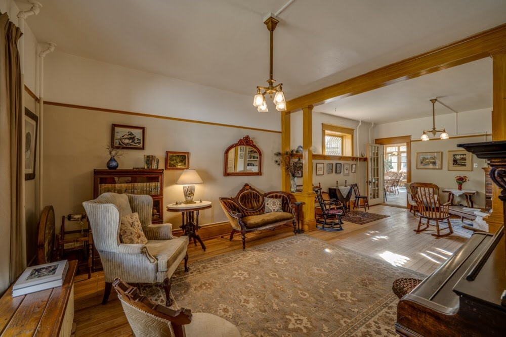 A spacious living area features elegant wooden accents and warm lighting from a chandelier. Vintage furniture, including a plush armchair and a small sofa, is arranged around a decorative rug. Pictures adorn the walls, adding character to the inviting space, which connects to a sunroom.