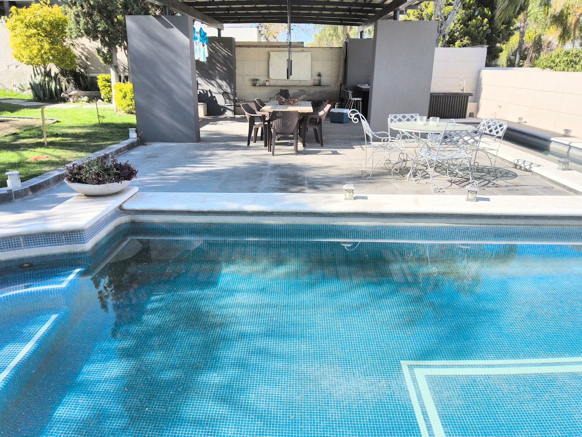 A private pool is framed by a spacious patio area shaded by a covered structure. The patio features a large dining table and several chairs, with a grill nearby. Lush greenery and garden beds are visible in the background.