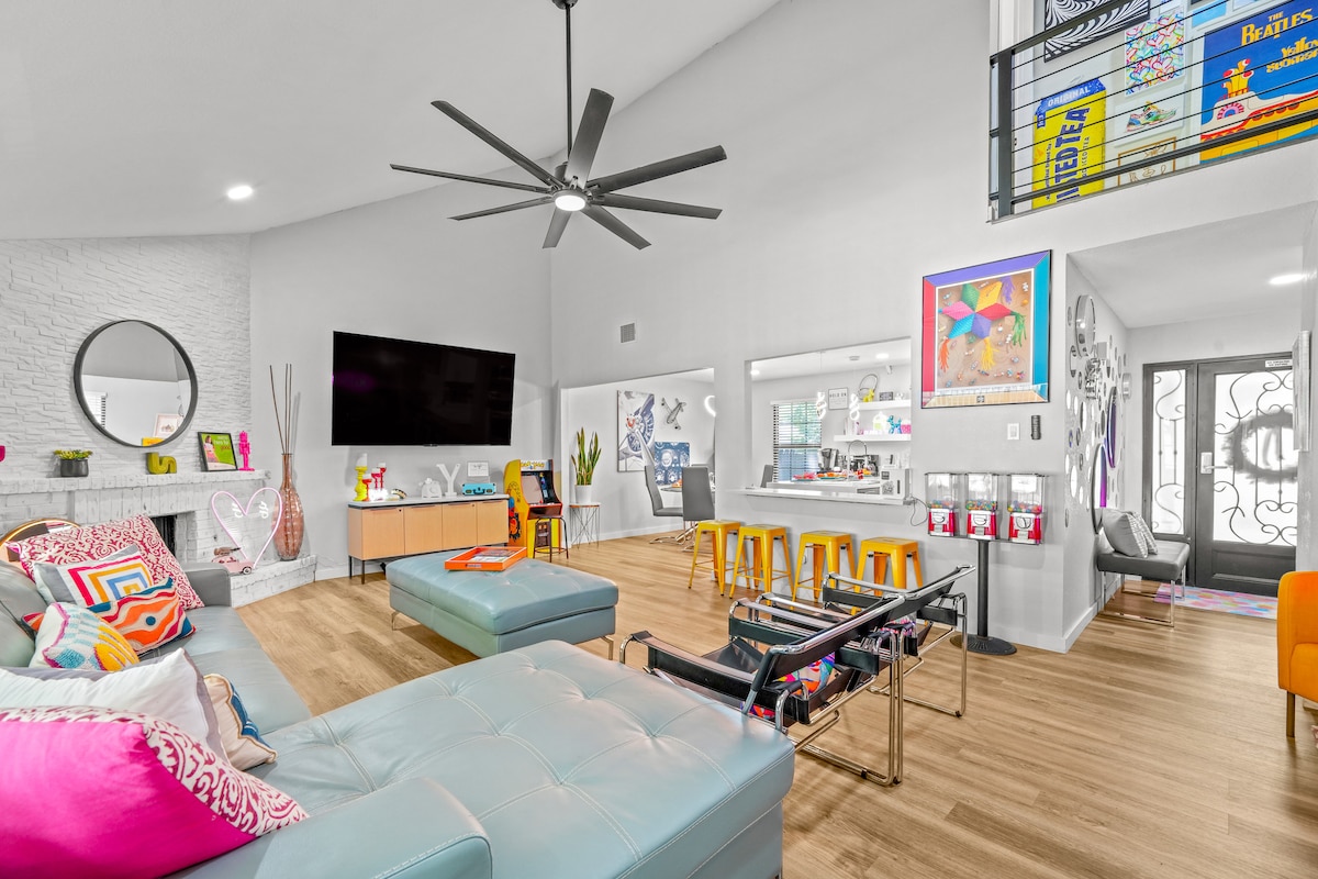 A modern living space features a large sectional couch adorned with colorful cushions. A flat-screen TV is mounted on the wall, opposite a bright kitchen area with yellow bar stools. Large windows allow natural light, complemented by a spacious layout that invites relaxation.