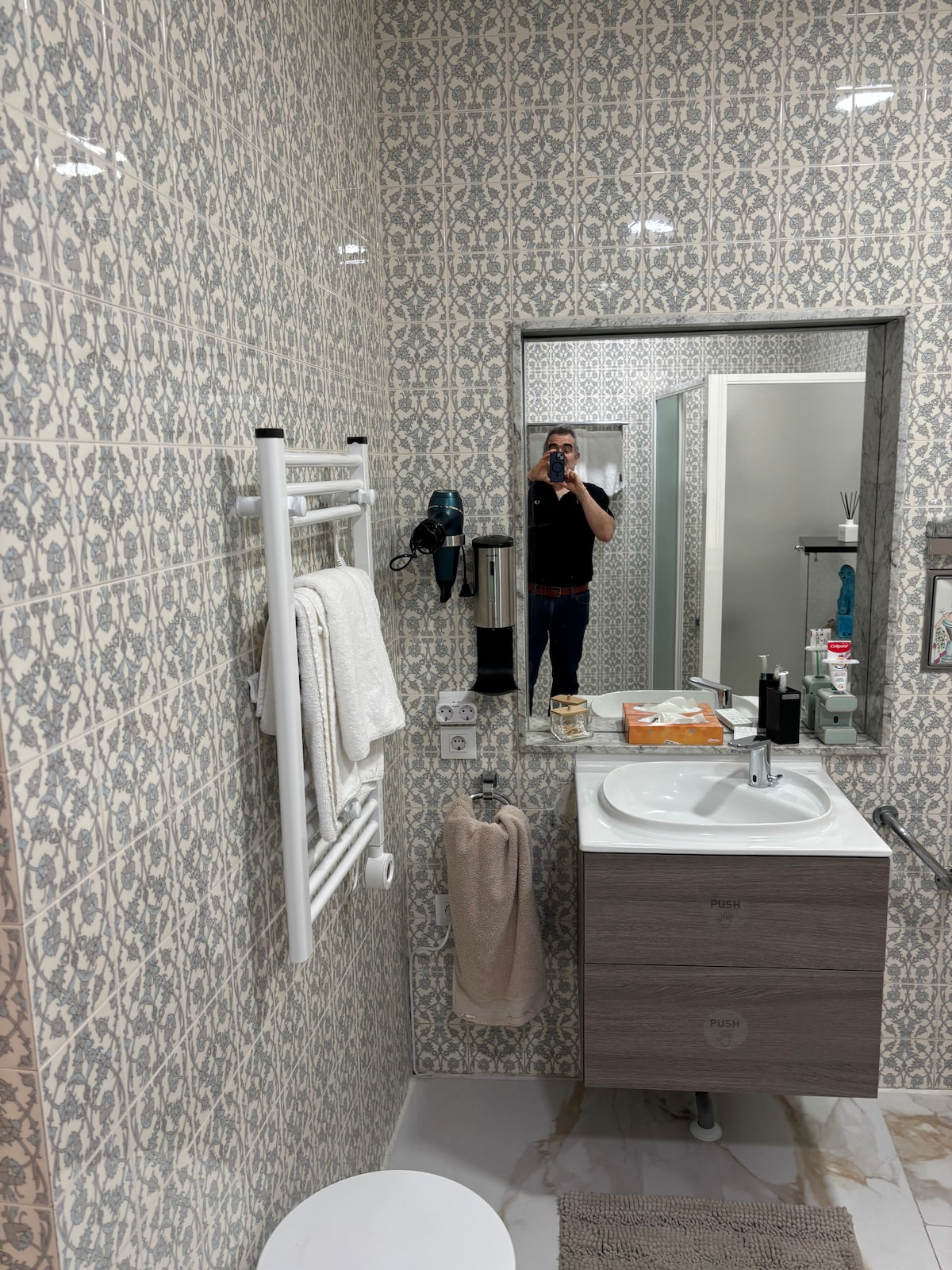 A modern bathroom is presented, featuring decorative patterned tiles on the walls. A clean sink with storage underneath is placed beside a large mirror. Towels are neatly hung on a rack, while various toiletries and a hairdryer are visible on the counter.