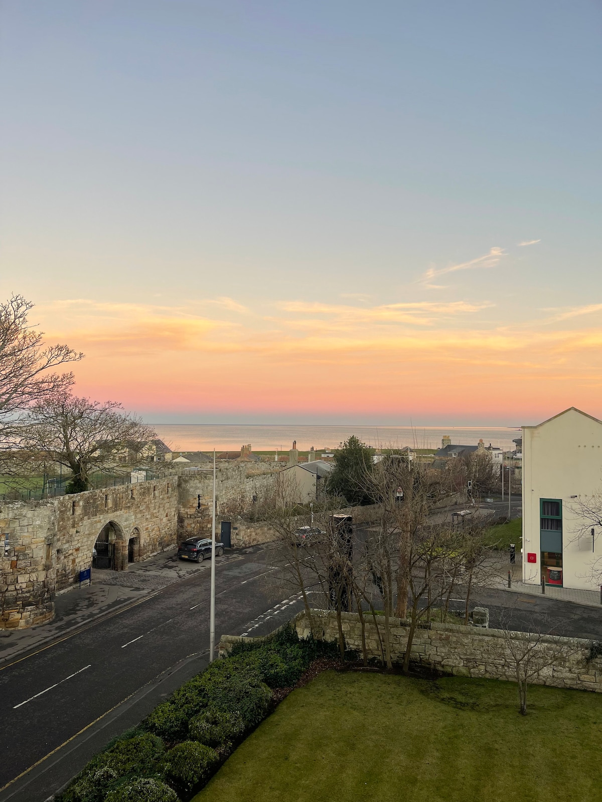 A serene view captures the horizon where the sea meets a pastel sky at dusk. A historic stone wall borders the street, with trees lining the road. The scene includes neatly manicured greenery in the foreground, creating a tranquil atmosphere.