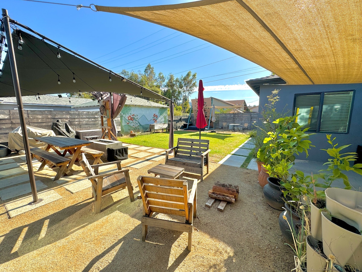 An outdoor space is presented with lounging areas under shade sails, complemented by wooden seating and a picnic table. Green grass is visible alongside stone paths, with a red umbrella providing additional shade. Various potted plants and a firewood stack enhance the inviting atmosphere.