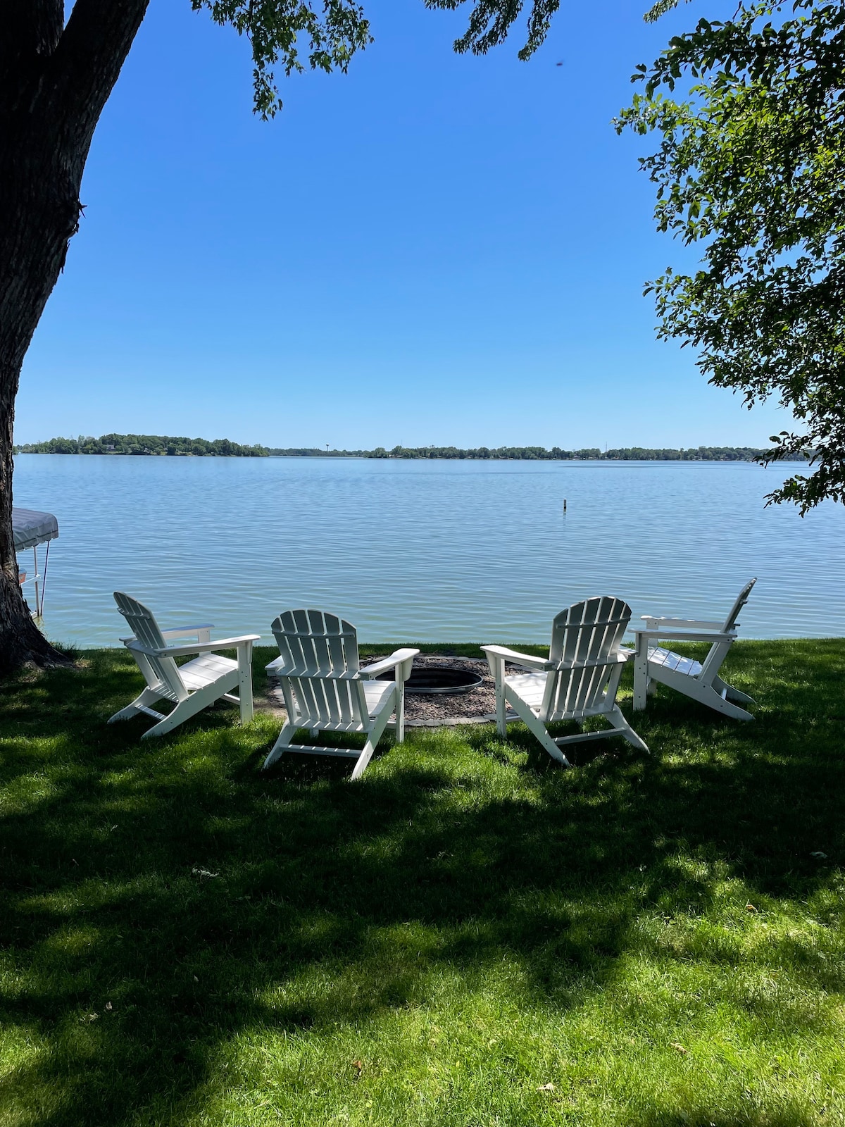 Four white Adirondack chairs are arranged in a semi-circle on a grassy area by the lake, overlooking calm water. A fire pit is centered among the chairs, with trees providing shade under a clear blue sky.