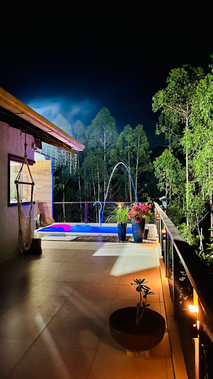 An outdoor area features a swimming pool illuminated with blue and purple lights, creating a serene atmosphere at night. Lush greenery surrounds the space, with potted plants and colorful flowers enhancing the view. A swing hangs nearby, inviting relaxation under the stars.