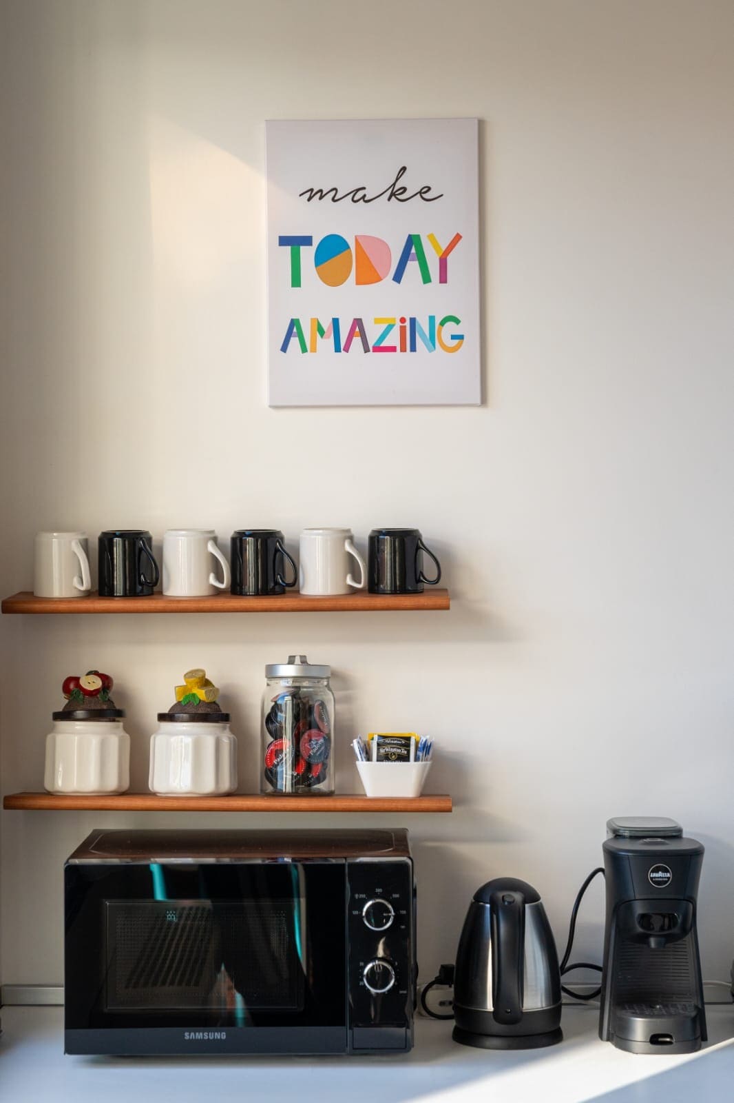 A kitchenette corner features a microwave and coffee machine alongside a kettle. Shelves display an assortment of mugs and containers, while a colorful wall art piece with the phrase 'make TODAY AMAZING' adds a cheerful touch.