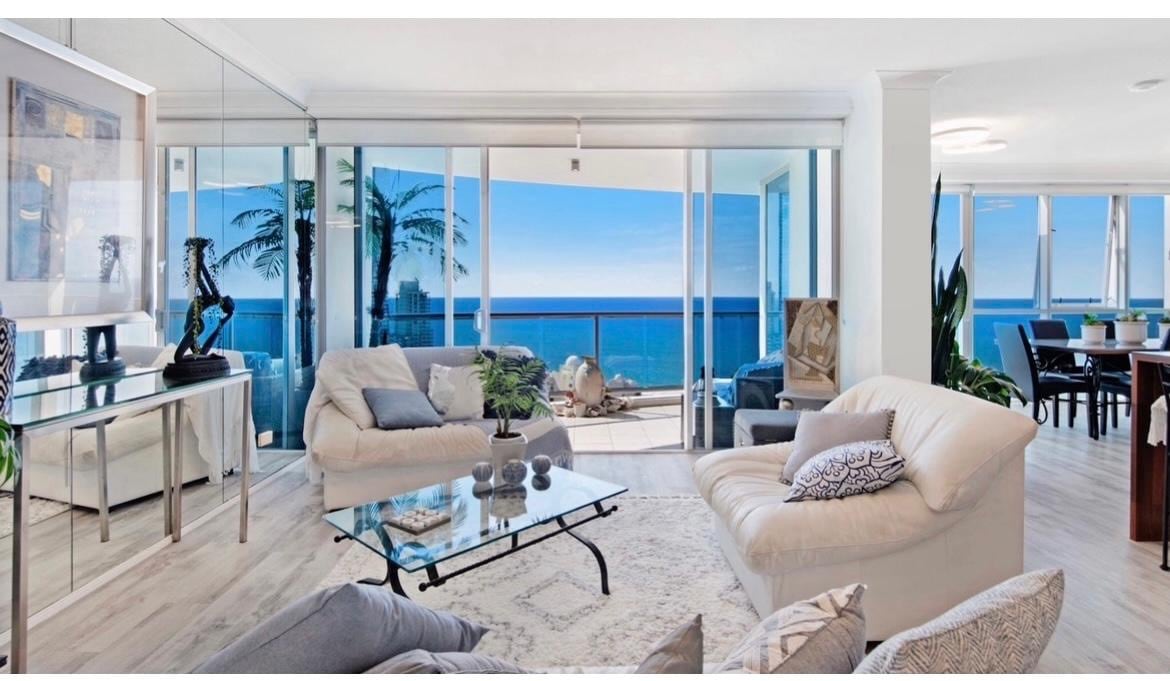 A bright and airy open-plan living area features expansive floor-to-ceiling windows, offering panoramic ocean views. Modern furniture, including plush sofas and a glass coffee table, is arranged around a light-colored rug. A spacious dining area is visible in the background, complementing the elegant decor.