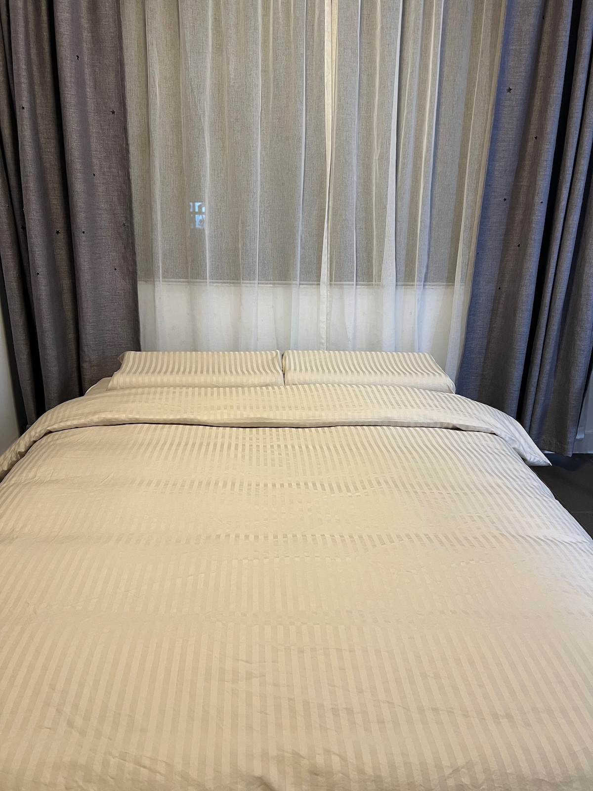 The sleeping area features a spacious bed dressed in soft, striped bedding. Sheer curtains frame a window, allowing natural light to filter through while providing a sense of privacy. The minimalist decor creates a calm and restful environment.