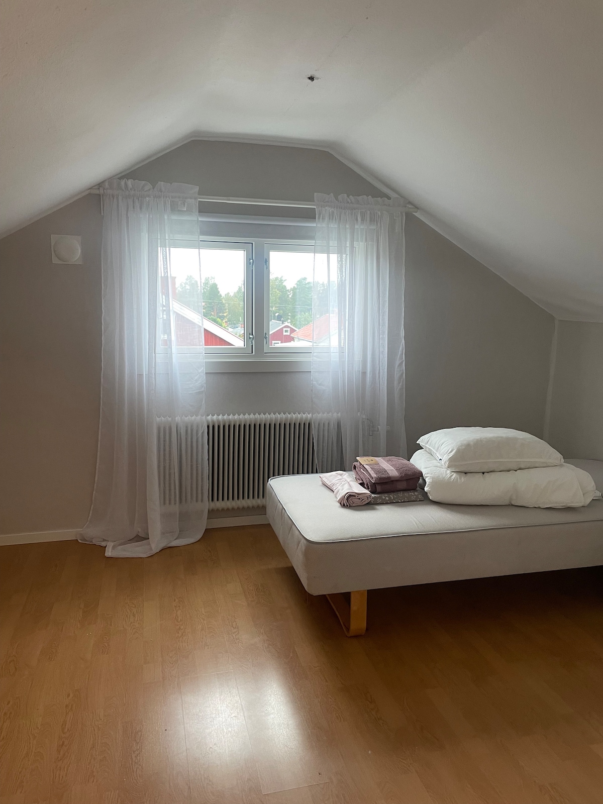 A simple sleeping area features a low-profile bed with neatly arranged pillows and blankets. Natural light filters through a large window dressed with sheer curtains, illuminating the light-colored walls and wooden flooring. A radiator is positioned beneath the window.