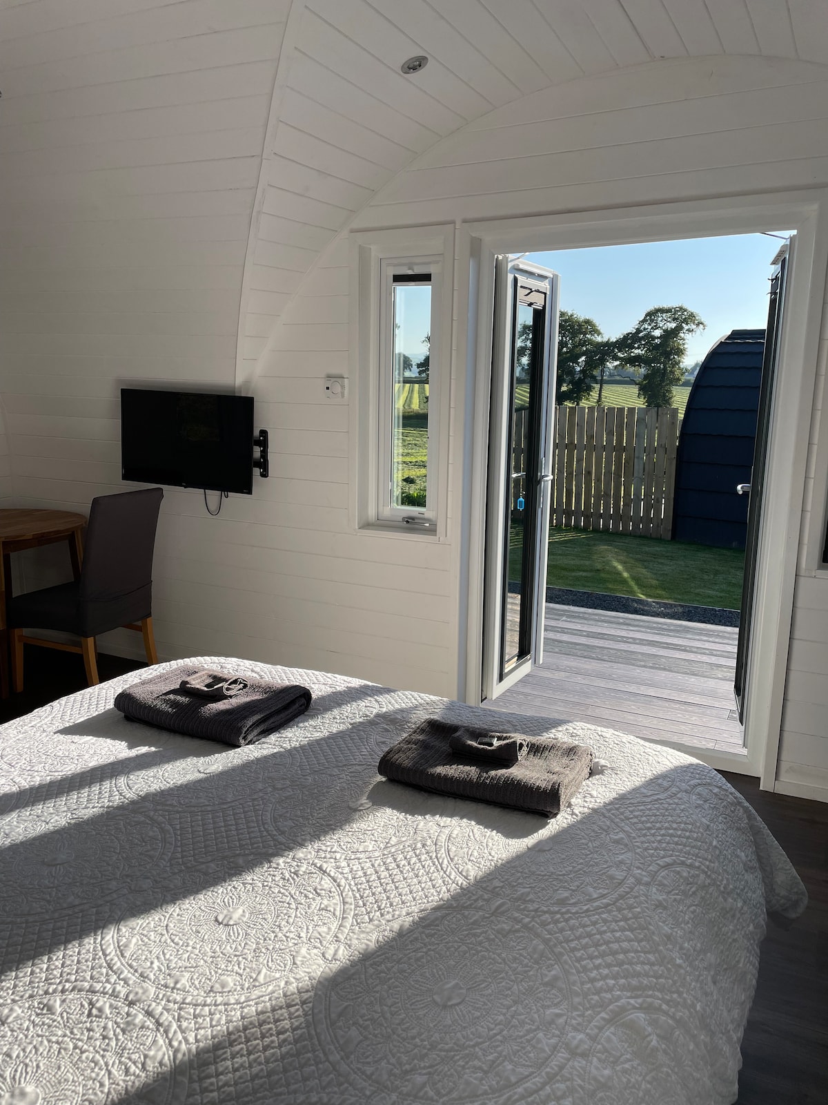 A spacious sleeping area features a neatly made bed with two towels placed decoratively on top. Natural light floods the room through an open door, leading to a private deck. A small dining table is positioned nearby, with a wall-mounted TV visible.