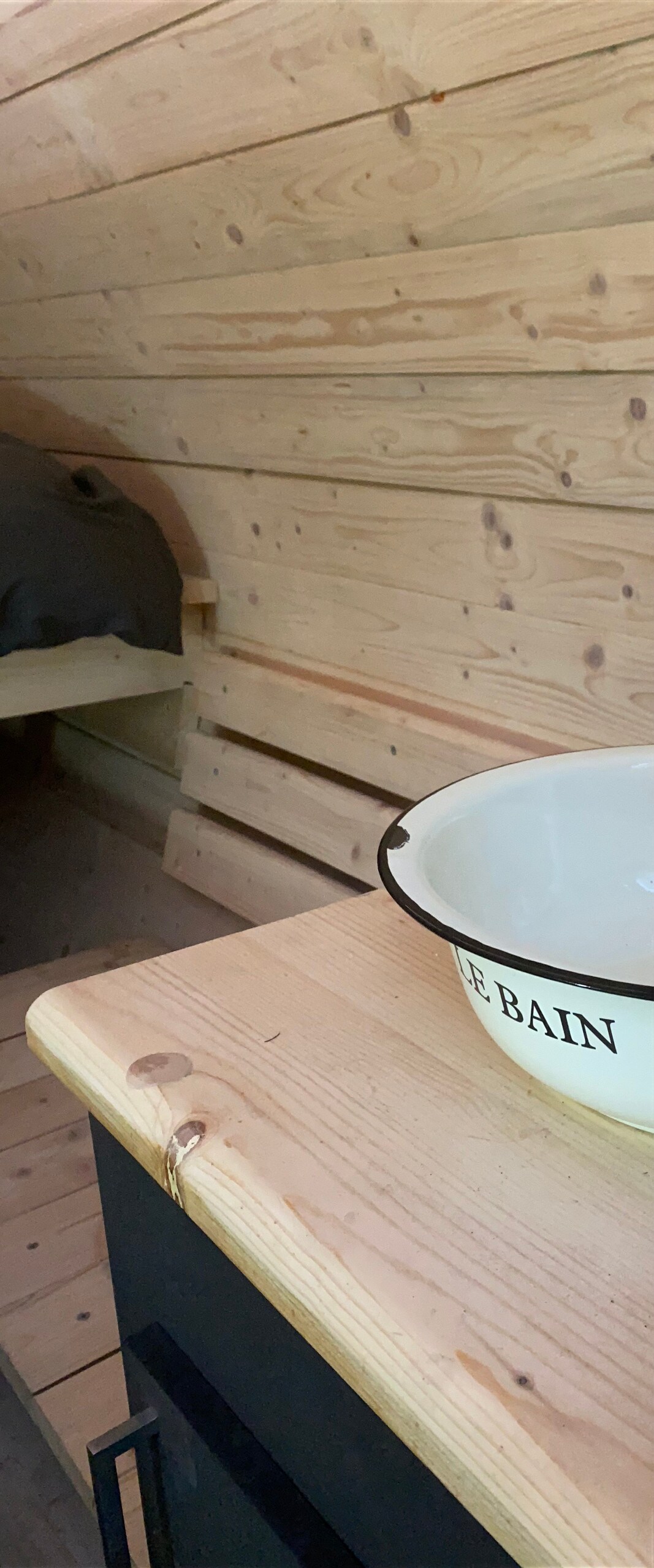 A wooden interior showcases light wooden walls and furnishings, creating a cozy ambiance. A bowl labeled 'LE BAIN' rests on a table, adding a rustic touch. A cozy sleeping area, defined by a simple bed, is positioned in the background.