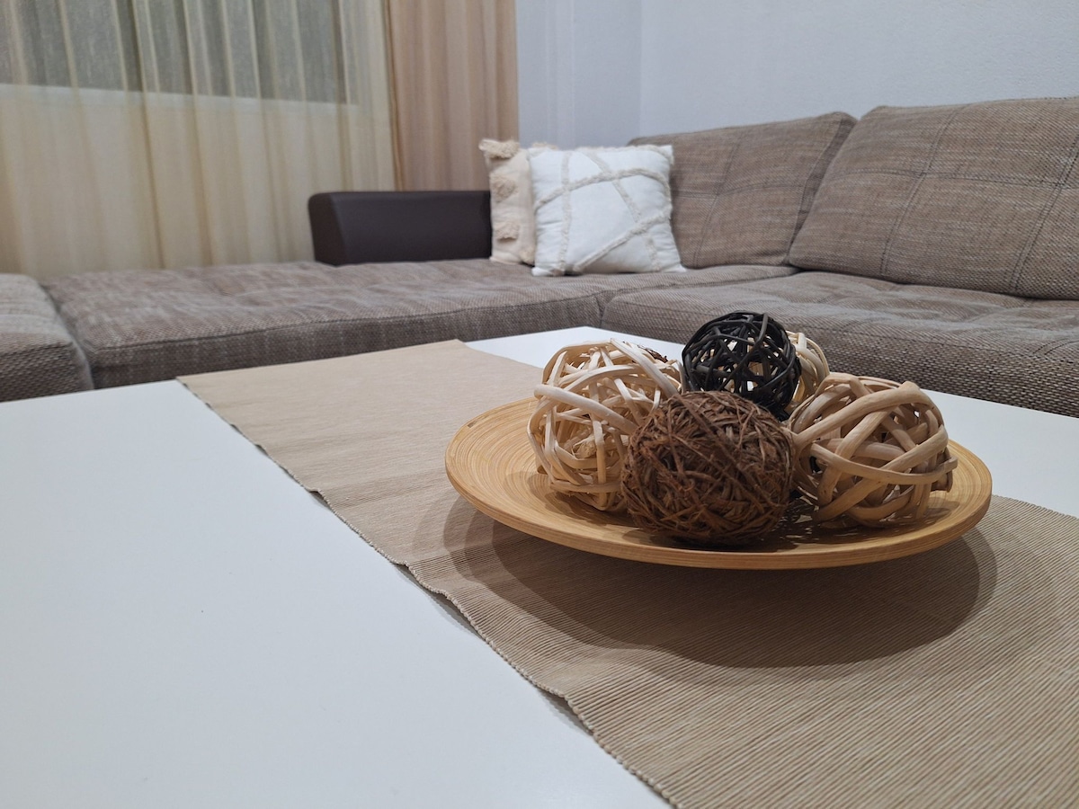 A spacious living area features a comfortable couch with neutral tones and a decorative throw pillow. A light-colored table is set with a circular platter displaying natural decorative balls, adding a touch of texture to the space.