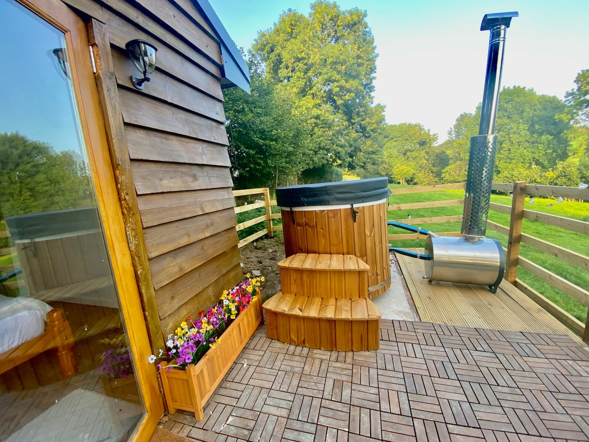 An outdoor deck showcases a wooden hot tub surrounded by a low wooden barrier and stairs leading to it. A planter with colorful flowers adds a touch of vibrancy. The structure is complemented by a chimney, indicating a log-fired heating system.