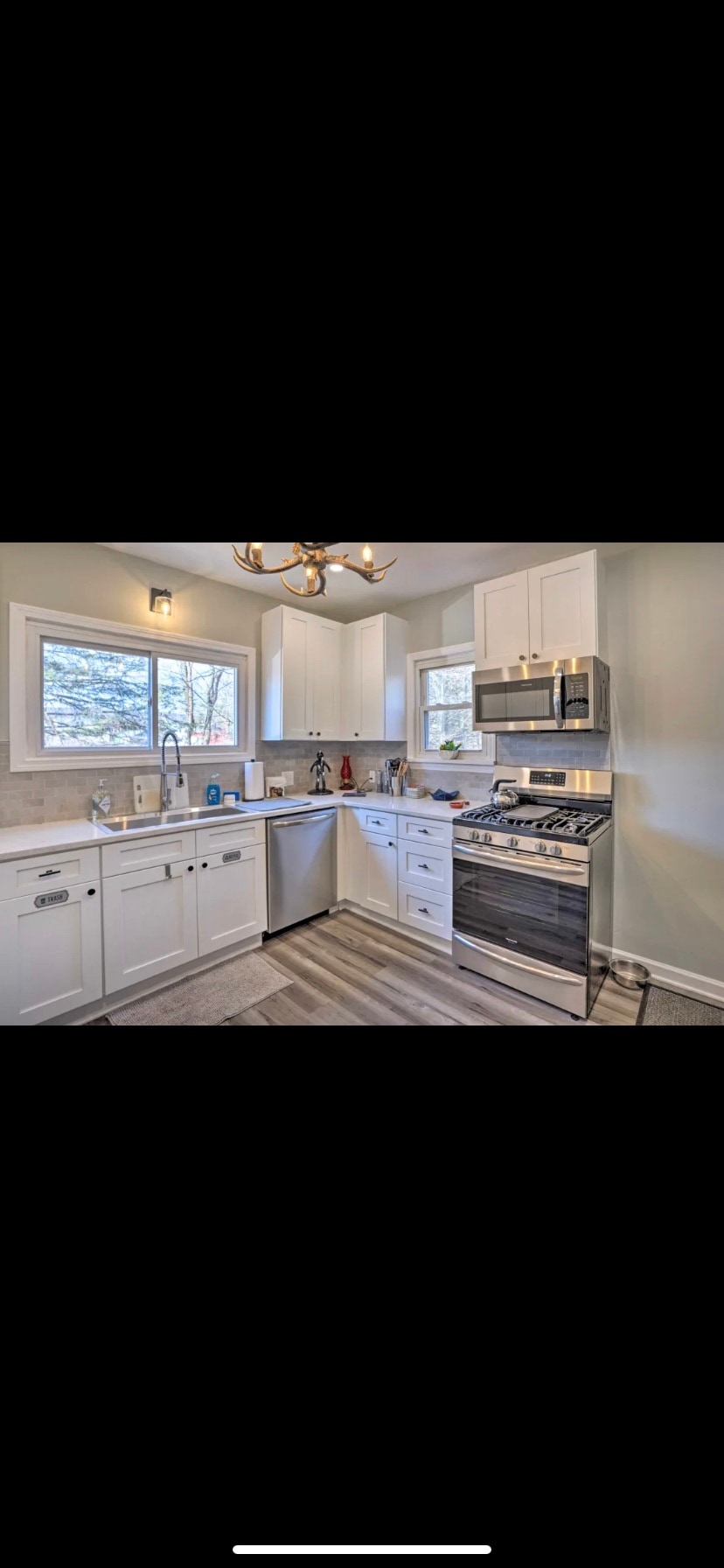A well-equipped kitchen features white cabinetry and stainless steel appliances. Two windows allow natural light to enter the space. A countertop with a modern sink is positioned next to a stove and oven. The flooring consists of light-colored planks, adding to the bright atmosphere.