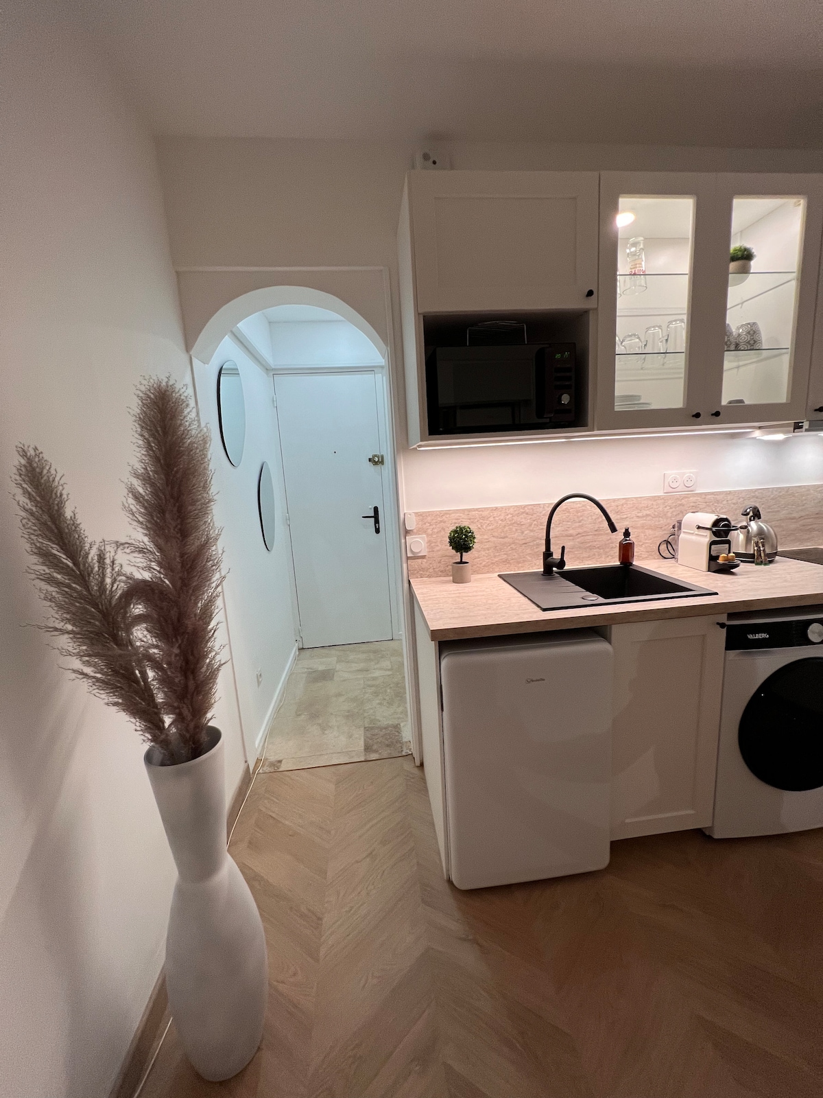 The image showcases a modern kitchenette with sleek cabinetry and a countertop. A compact sink and a small refrigerator are visible. A washer is integrated into the cabinetry. A decorative white vase with pampas grass adds a touch of greenery to the space, enhancing the overall aesthetic.