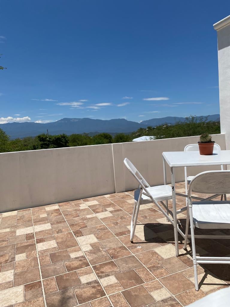 An outdoor terrace features a light-colored tiled floor with a simple white table and four chairs. A small potted plant is placed on the table, offering a view of distant mountains against a clear blue sky.