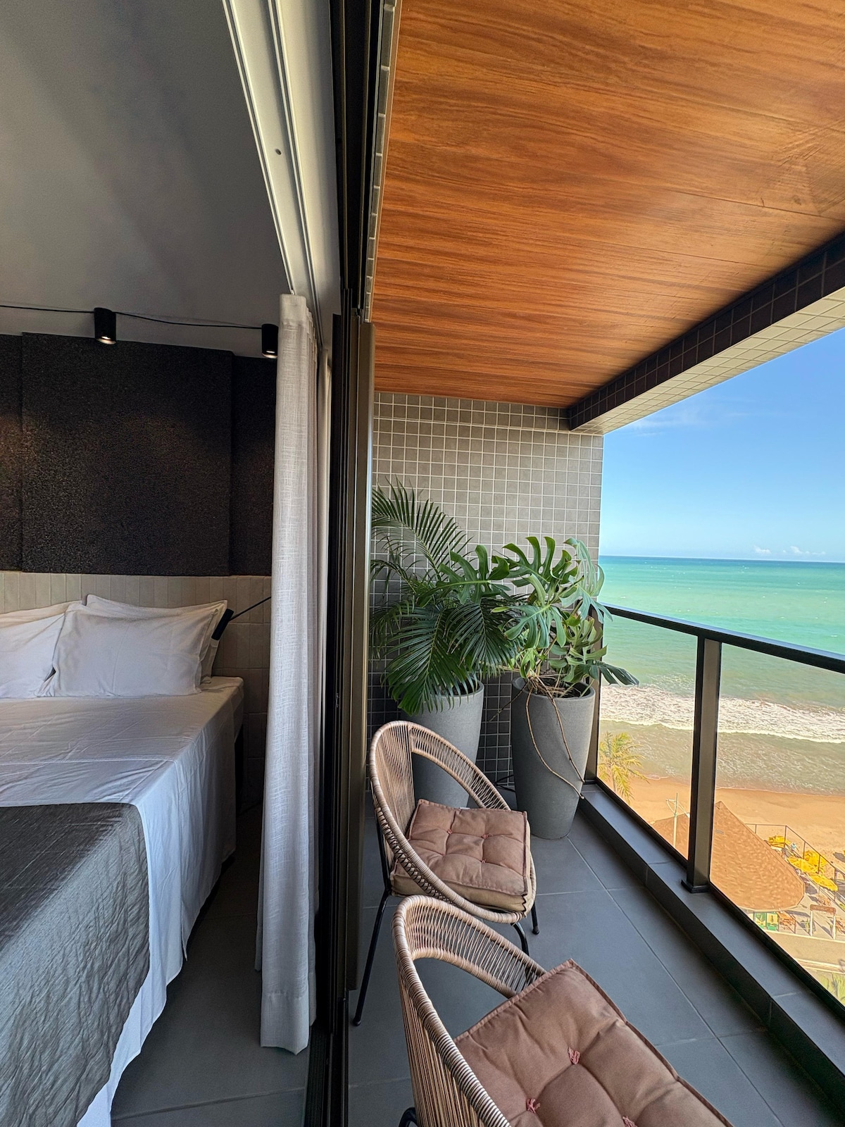 A spacious bedroom is visible to the left, featuring a neatly made bed with white linens. On the right, a private balcony offers views of the ocean, complemented by two wicker chairs and potted plants. The warm wood ceiling adds a touch of comfort.