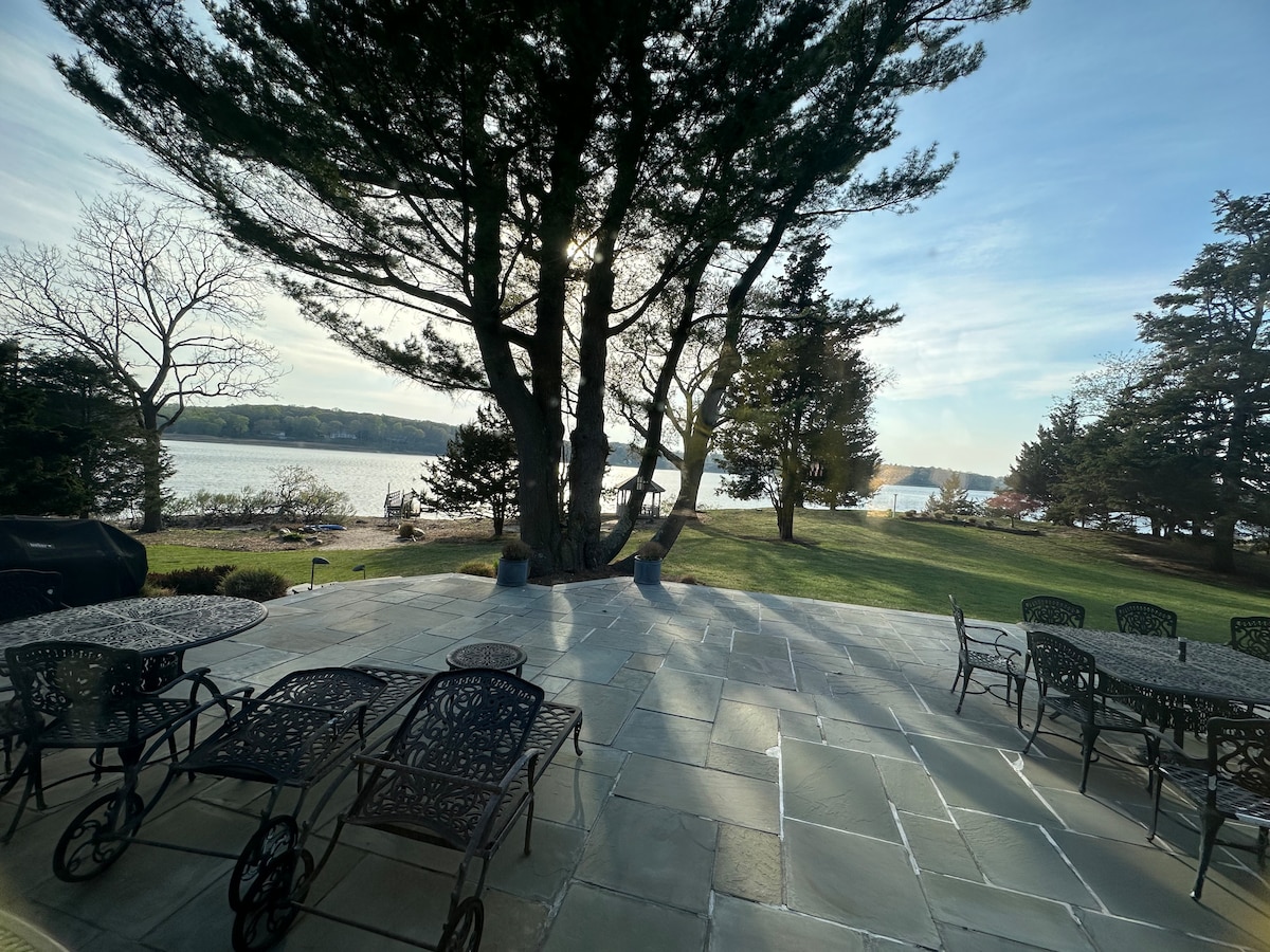 A stone patio is featured, furnished with multiple wrought iron tables and chairs. The space overlooks a tranquil waterfront, framed by the shadows of tall trees, offering a serene outdoor area for relaxation.