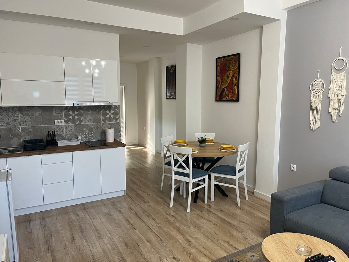 A modern kitchen and dining area are showcased, featuring a white cabinetry and brown countertop. A round table is set with four chairs, complemented by decorative wall art. The spacious living area includes a soft gray sofa, creating an inviting atmosphere for relaxation.