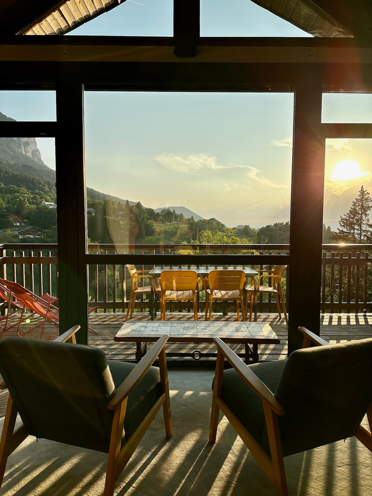 Large glass doors frame a serene view of the surrounding mountains and valley. A spacious balcony is visible, furnished with a dining table and chairs. Two comfortable armchairs sit in the foreground, inviting relaxation while enjoying the sunset.