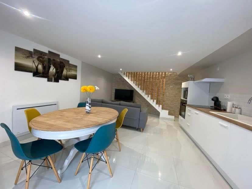 A modern living space features a round wooden dining table surrounded by colorful chairs. A soft gray sofa is positioned nearby, and a television is mounted on the wall. A staircase leads to the upper level, while recessed lighting enhances the bright atmosphere.