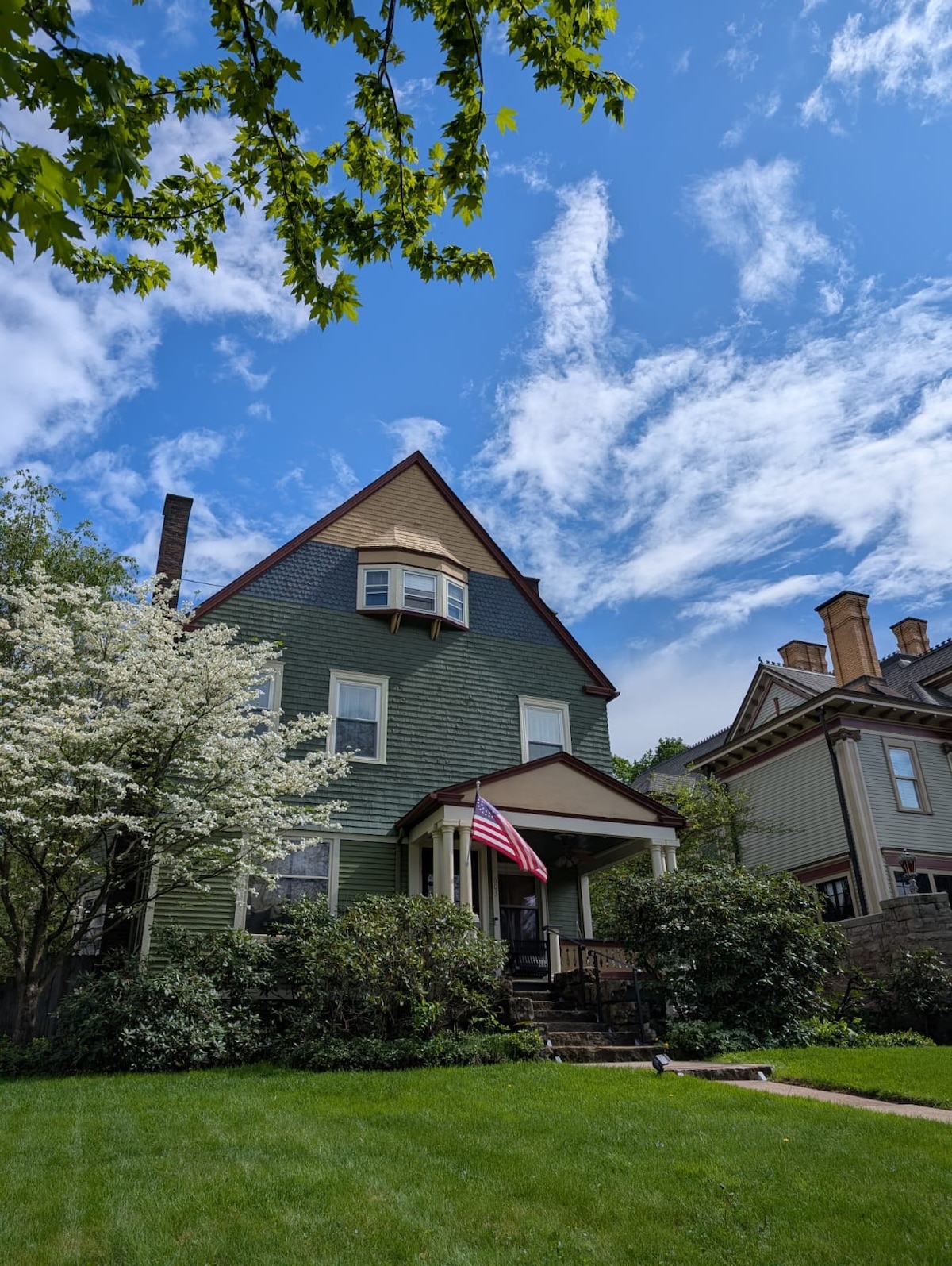 A historic Victorian home is presented with a unique triangular roof and green exterior. Flanking trees and flowering shrubs add to the charm. An American flag is displayed at the front, welcoming guests. Clear blue skies and scattered clouds enhance the home's inviting presence.