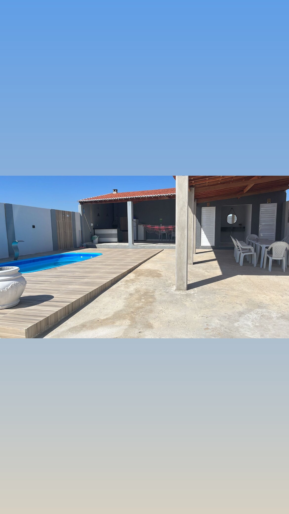An inviting outdoor space is highlighted by a swimming pool, bordered by wooden decking. A covered patio area features a dining setup with white chairs, and a shaded area offers views of the surrounding landscape.