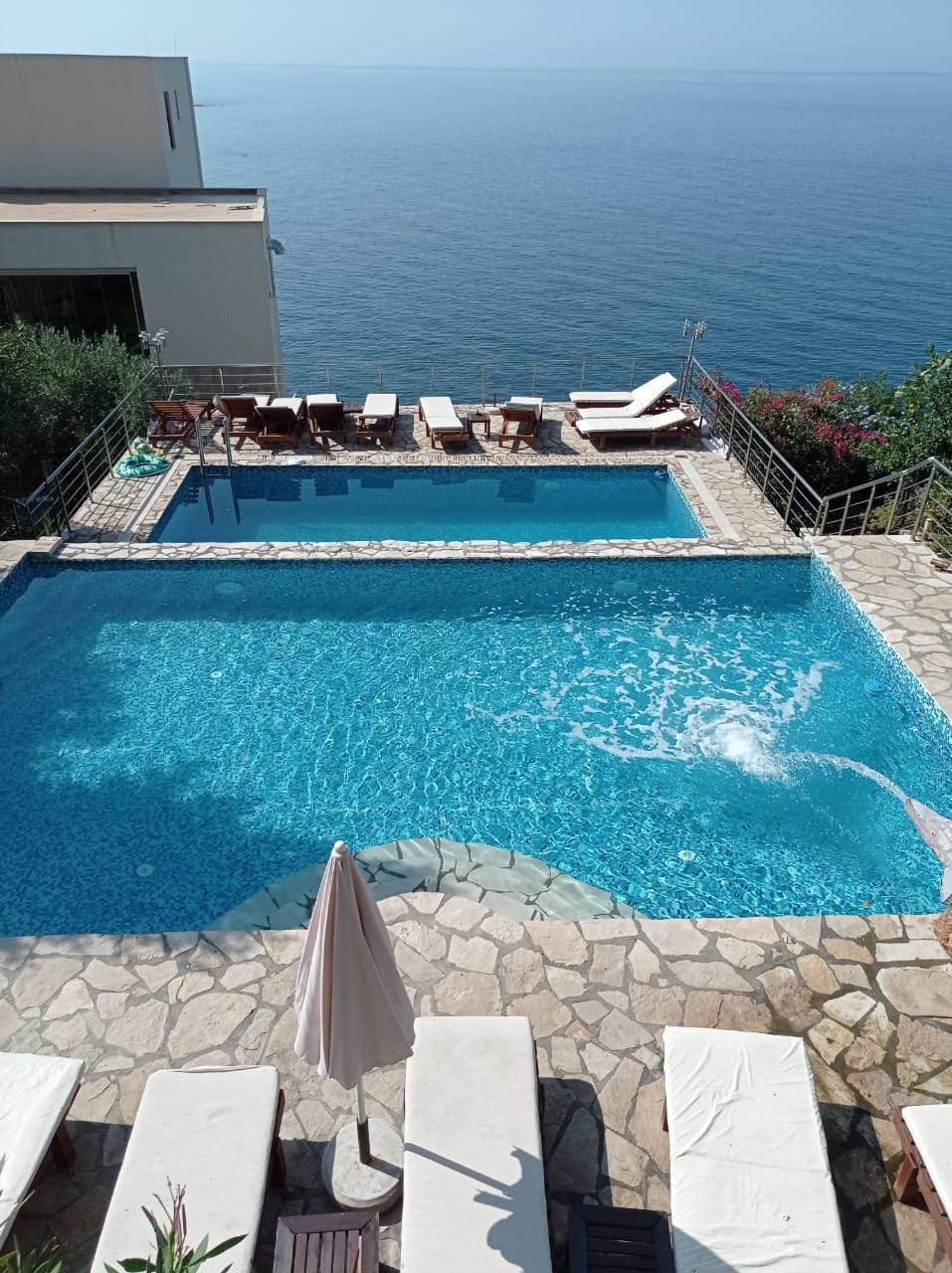 A spacious swimming area features two pools, one with a gentle current, and is surrounded by a natural stone deck. Lounge chairs are arranged nearby, offering a view of the calm sea and lush greenery along the edges.