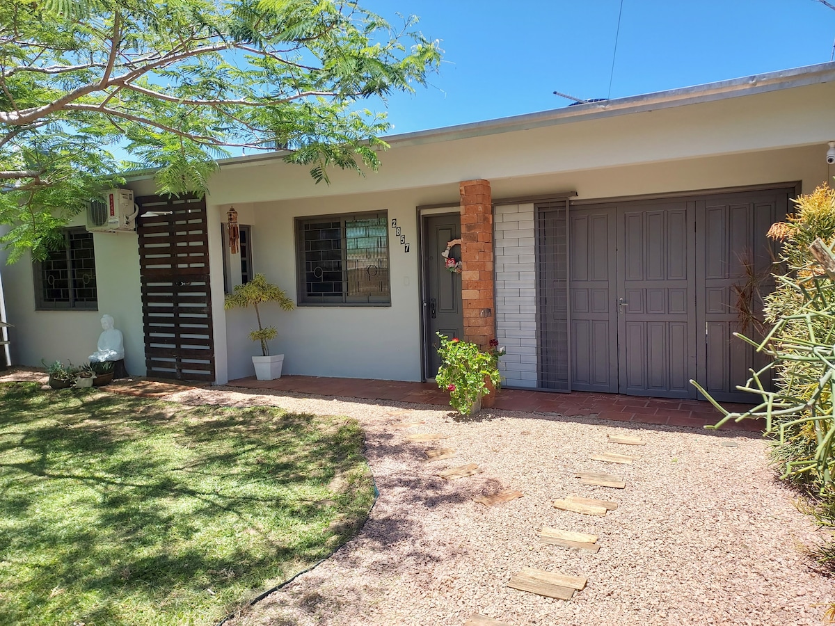The exterior of a family-friendly house is shown, featuring a well-maintained garden with a variety of green plants. The entrance includes a combination of brick and neutral-colored walls, flanked by secure doors. A shaded area is provided by a tree, enhancing the outdoor space.
