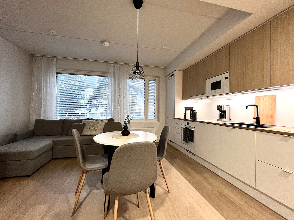 The open-plan living area features a comfortable sectional sofa and a circular dining table surrounded by four chairs. Natural light fills the space through a large window with sheer curtains, and a modern kitchen is equipped with essential appliances, creating a functional and inviting atmosphere.