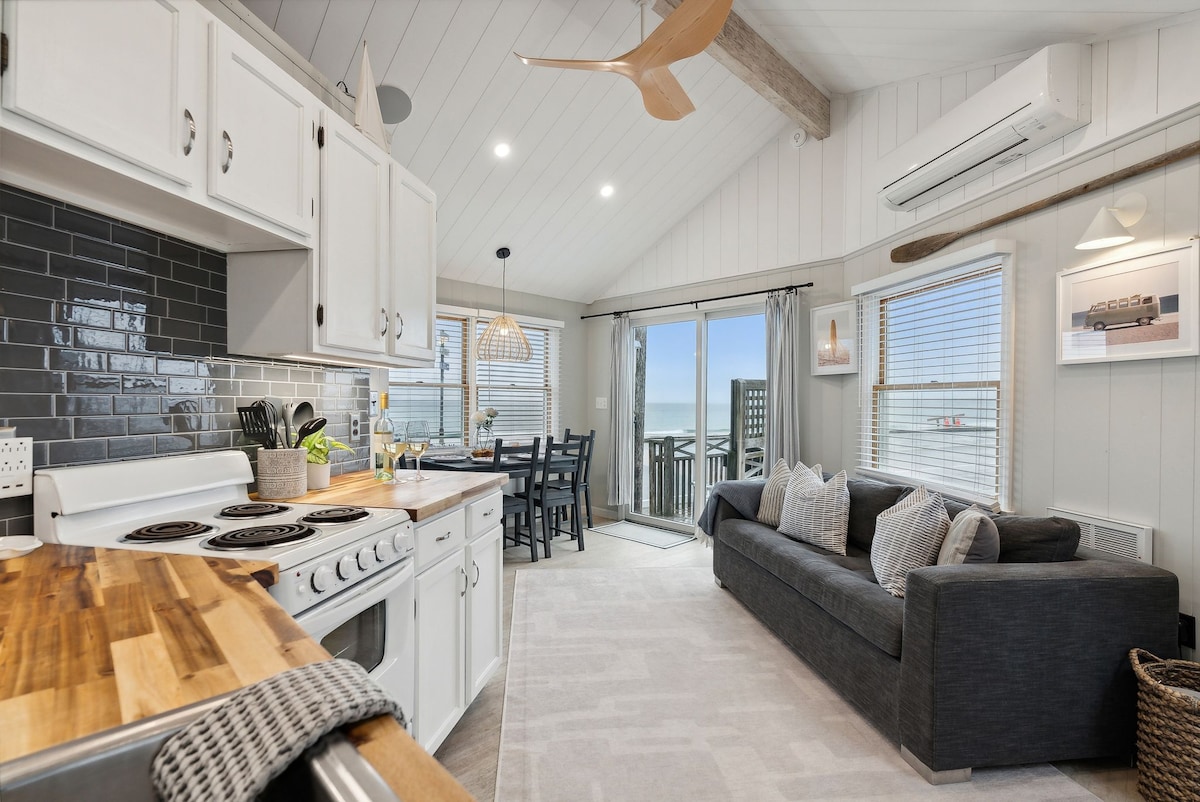 An open-concept living space features a well-equipped kitchen with light cabinetry and a wooden countertop. A cozy seating area is adjacent, with a dark sofa, and natural light streams through large windows, offering views of the ocean. A dining table is positioned nearby.