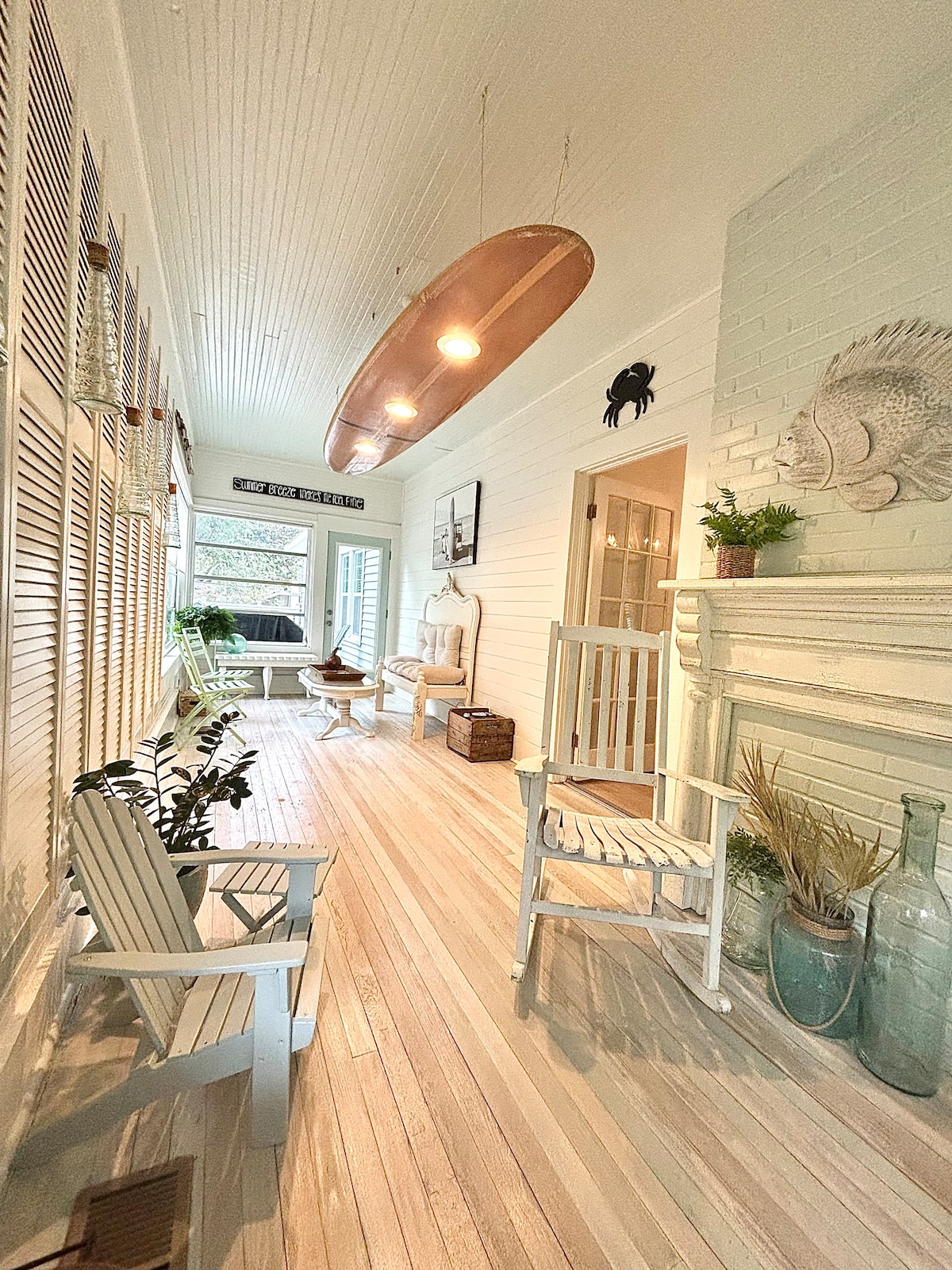A sunlit sunroom features light wooden flooring and a row of windows, allowing natural light to fill the space. Two white chairs are positioned along the walls, accompanied by decorative plants and coastal-inspired accents, including a large copper surfboard hanging from the ceiling.