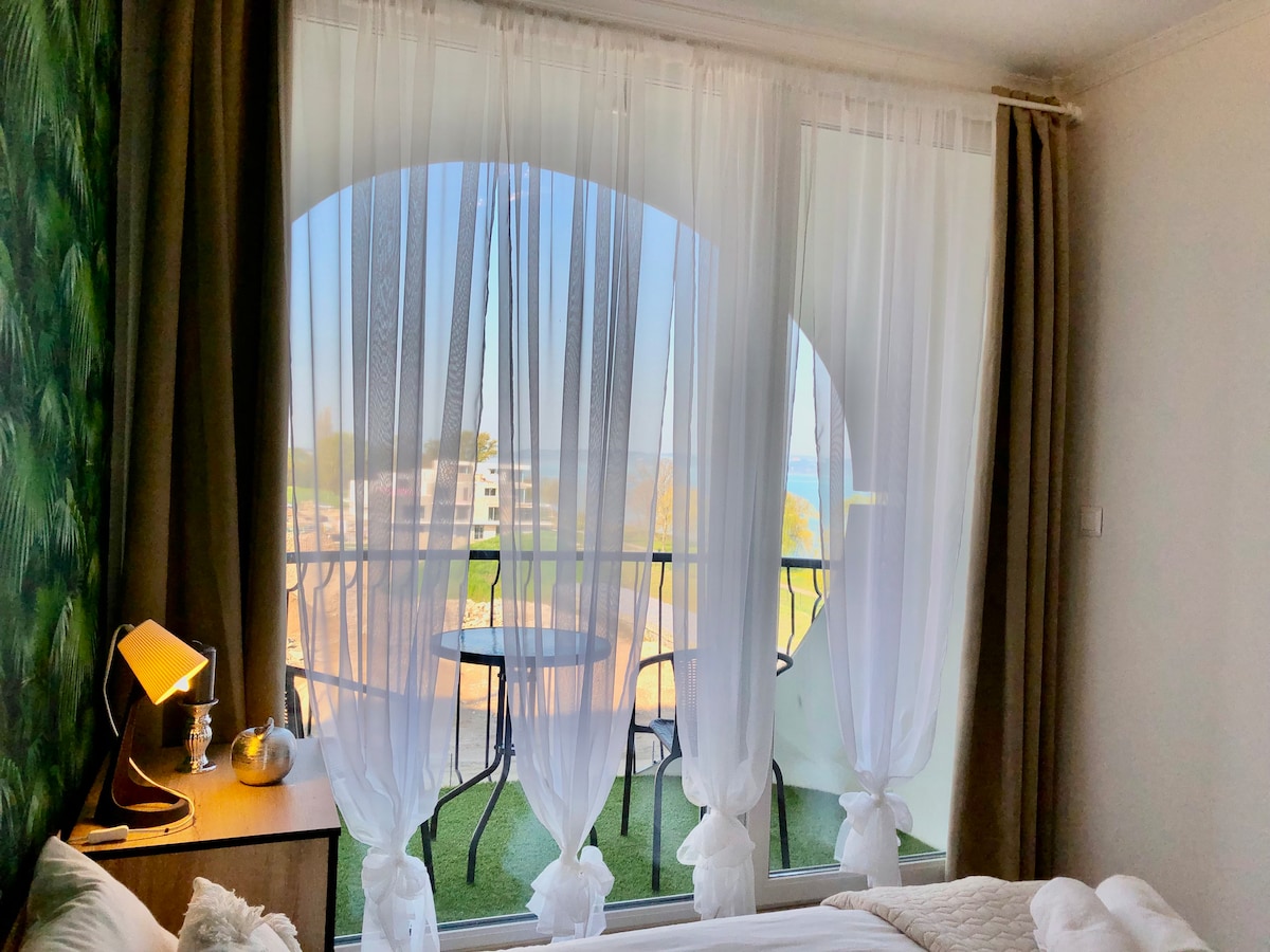 A bright interior is highlighted by sheer white curtains that frame a view of a balcony with a small table and chairs. Soft light filters through, revealing a glimpse of the surrounding landscape beyond.