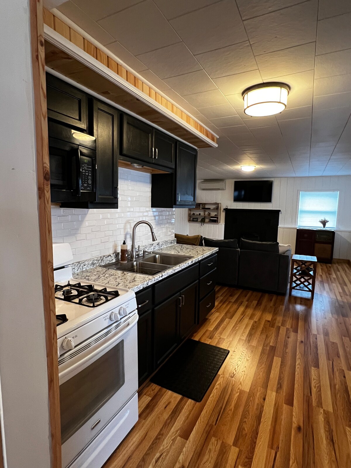 A fully equipped kitchen features dark cabinetry and a tiled backsplash. Appliances include a gas stove and a microwave. Hardwood flooring extends into the living area, which has a sofa and a flat-screen TV. Natural light enters through the window at the far end.