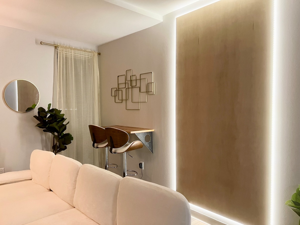 A well-lit living area is shown, featuring a soft, white sofa and a small dining bar with two sleek stools. A round mirror and decorative wall art enhance the serene atmosphere, while a plant adds a touch of greenery.