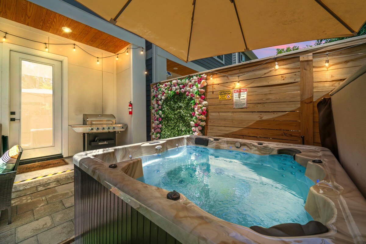 A private hot tub is set against a backdrop of vibrant flowers on a wooden wall. Soft lighting from overhead string lights enhances the outdoor space, while a grill is positioned nearby under an umbrella. The area is designed for relaxation and gatherings.