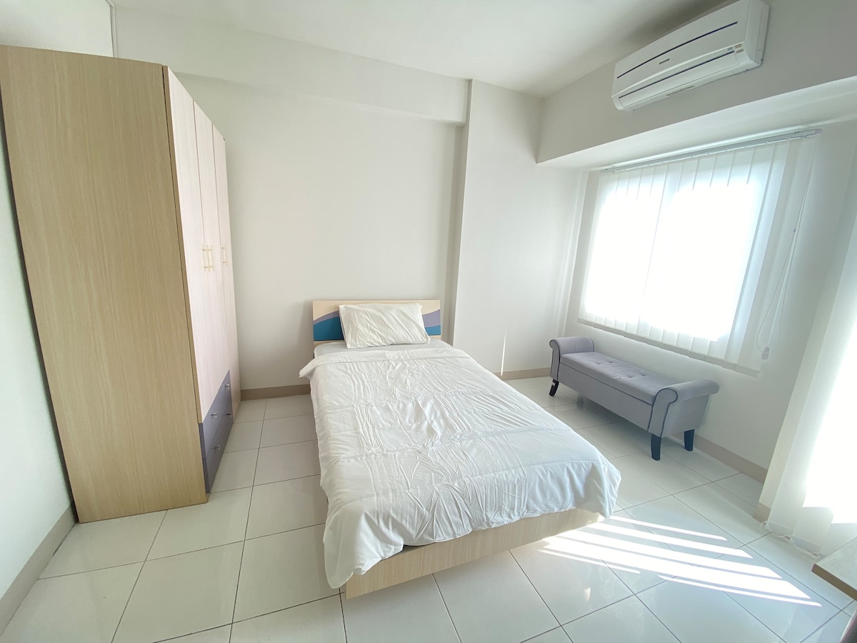 A bright sleeping area features a spacious bed with crisp white linens. Natural light filters through a large window with vertical blinds. A simple wardrobe and a small bench offer additional storage and seating options, creating an uncluttered and relaxing space.