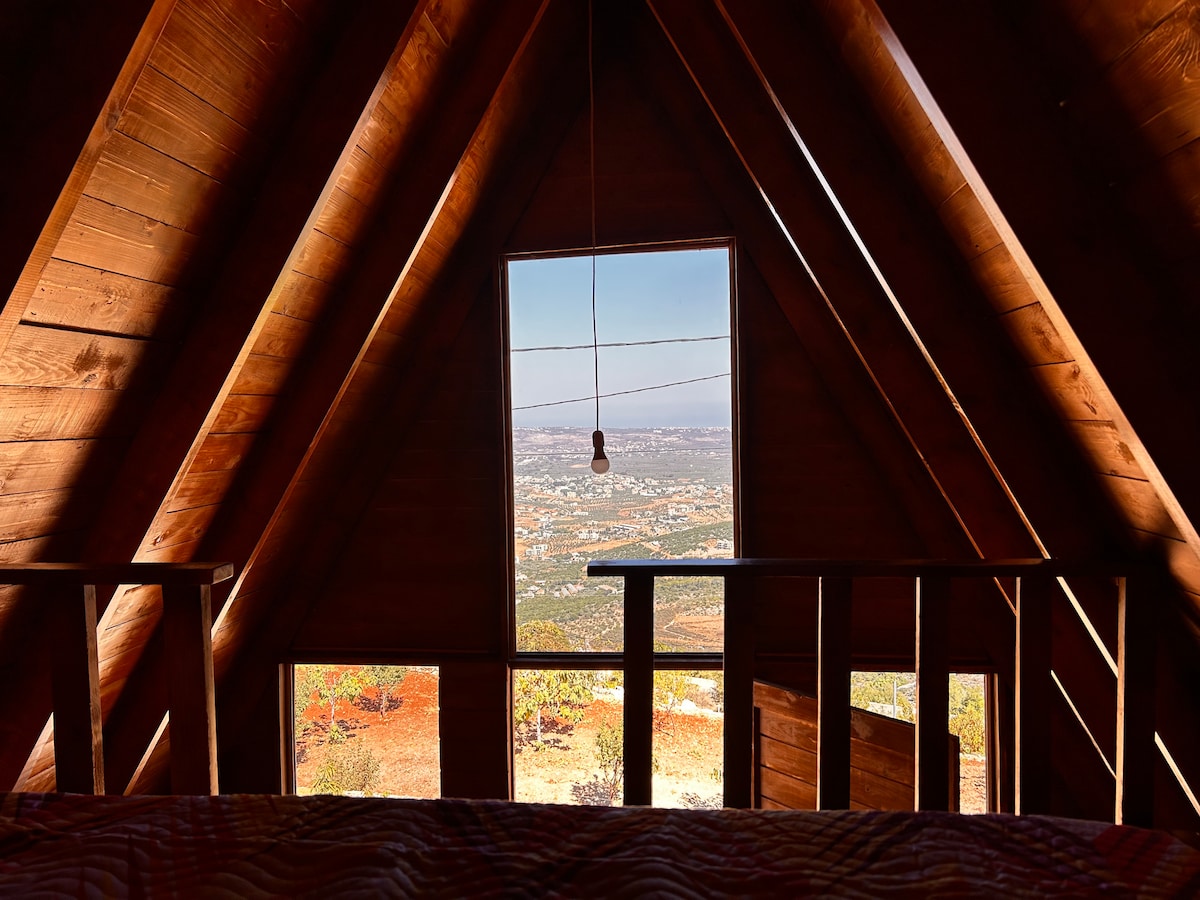 A view from inside a wooden cabin reveals large triangular windows showcasing a panoramic landscape. Natural wood finishes create a warm ambiance, while the surrounding greenery and distant hills provide a serene backdrop. A single pendant light fixture is suspended from the ceiling.