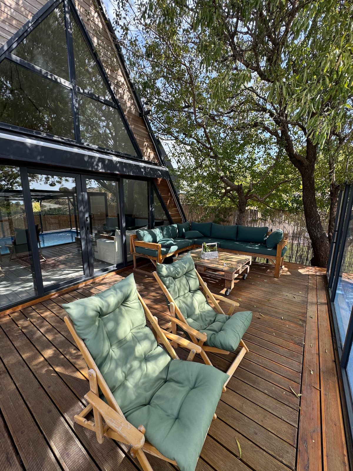 A spacious wooden deck is surrounded by greenery, featuring comfortable seating with plush green cushions. Large glass doors provide a view into the living area, while a blend of light and shadows from the trees enhances the outdoor setting.