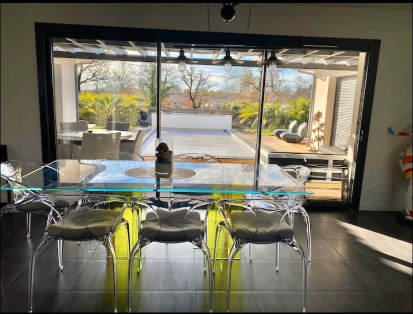 A dining area is shown with a sleek glass table surrounded by six transparent chairs. Behind, large sliding doors open to a spacious outdoor terrace adorned with lush greenery and comfortable seating options. Natural light enhances the contemporary design of the space.