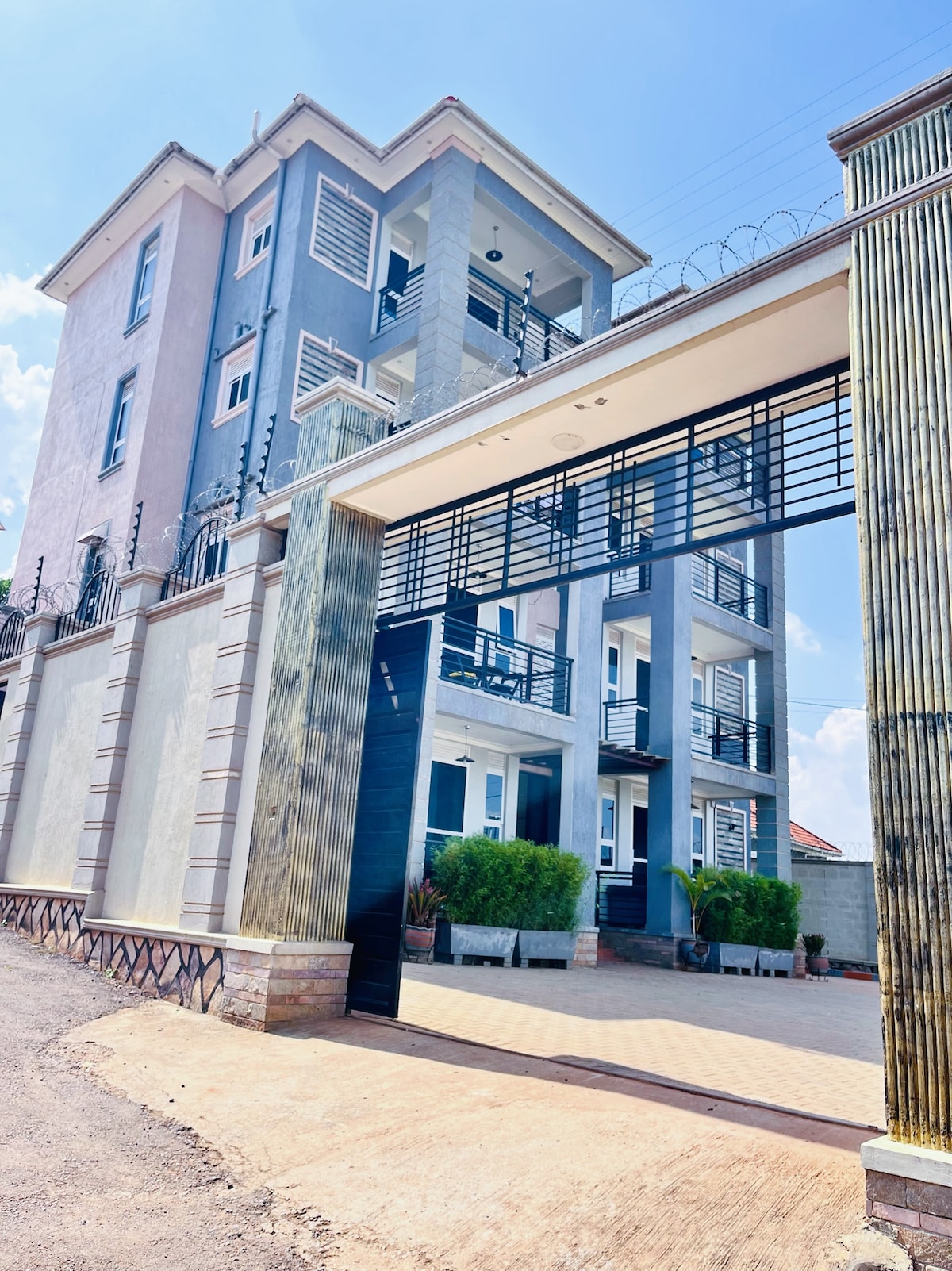 A modern three-story building is framed by a large gated entrance. Lush greenery is visible near the entrance, adding to the building's overall aesthetic. The structure features multiple balconies with doors, enhancing the sense of openness and connection to the outdoors.