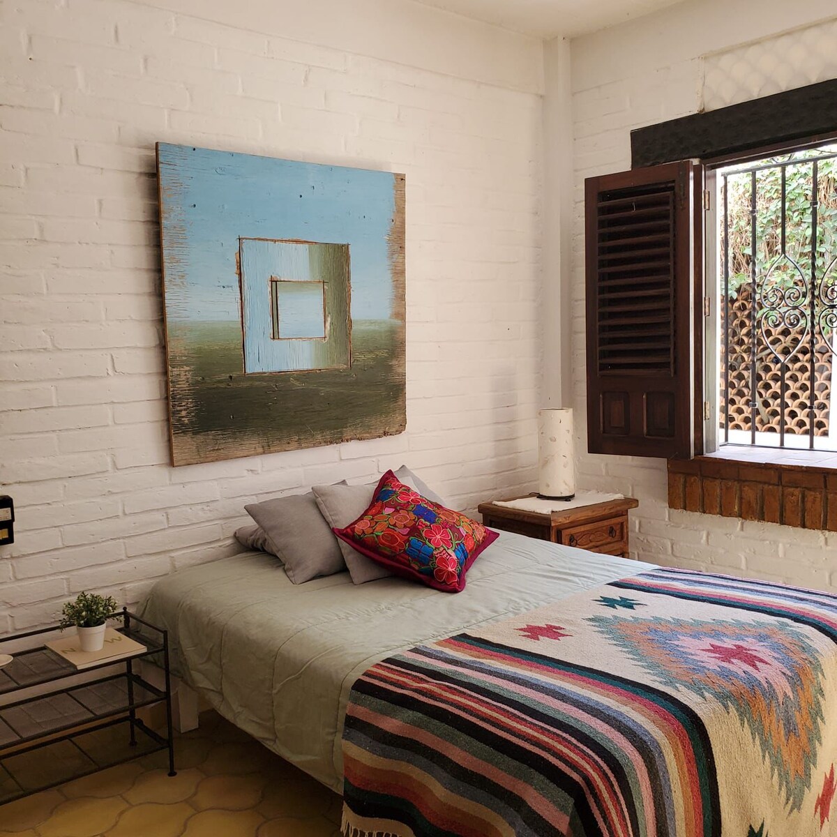 A cozy sleeping area features a spacious bed covered with a colorful woven blanket. A vibrant artwork is displayed above the bed. Natural light enters through a window adorned with wooden shutters, illuminating the room's white brick walls and wooden accents.