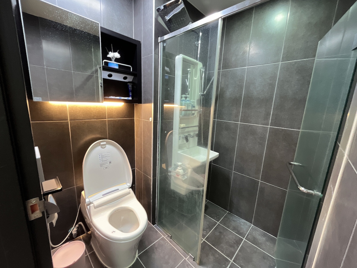 A modern bathroom features sleek black tiled walls and floor, with a glass shower enclosure on one side. A wall-mounted sink is positioned beside the shower, and a contemporary toilet is located opposite. Soft lighting enhances the clean lines and minimalist design.